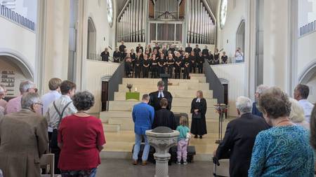 Pfarrer Garleff segnet Pfarrerin Therese Wagner mit Ehemann und Tochter am Taufbecken. Die Gemeinde sieht zu. Im Hintergrund steht der Chor.